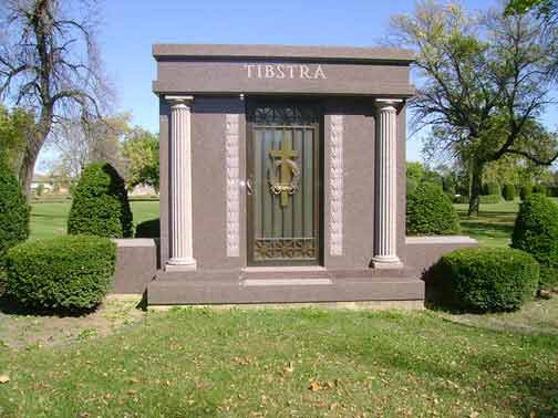 Tomb with the name Tibstra. Dark brown with pillars, a gold door with cross. Green lawn and bushes.