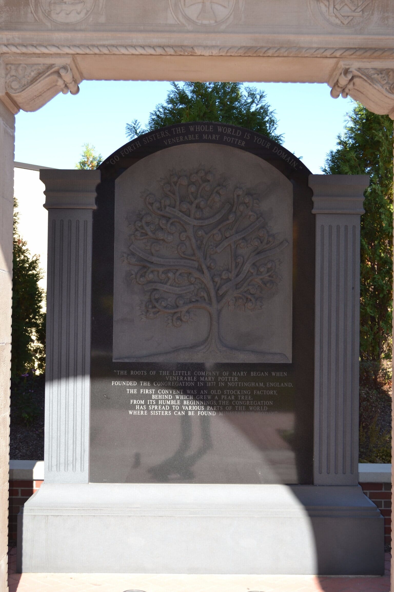 Stone memorial with a carved tree and inscription, framed by columns, set in a brick courtyard.