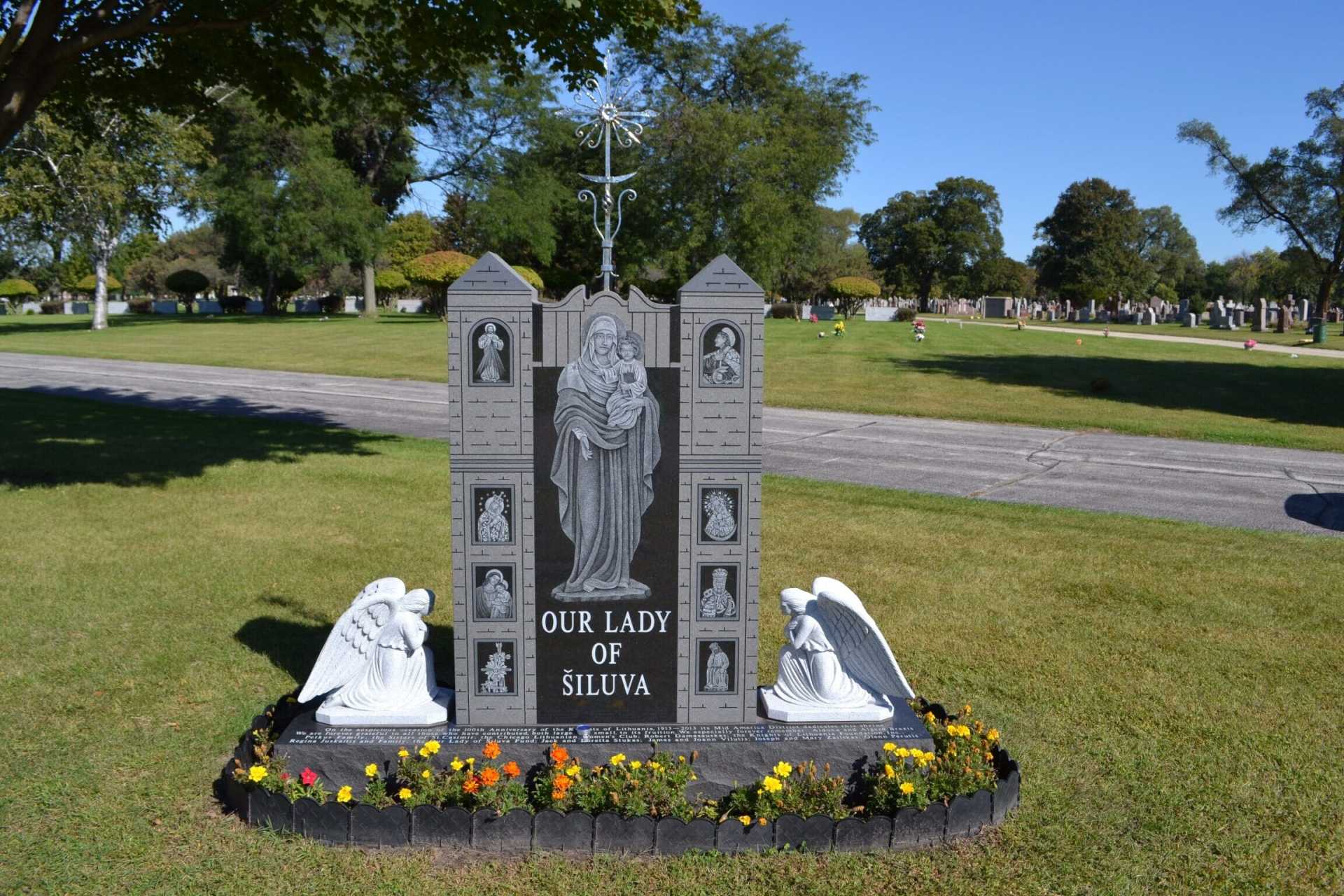 A large gray monument with a depiction of Our Lady of Silva in a cemetery, flanked by angels.