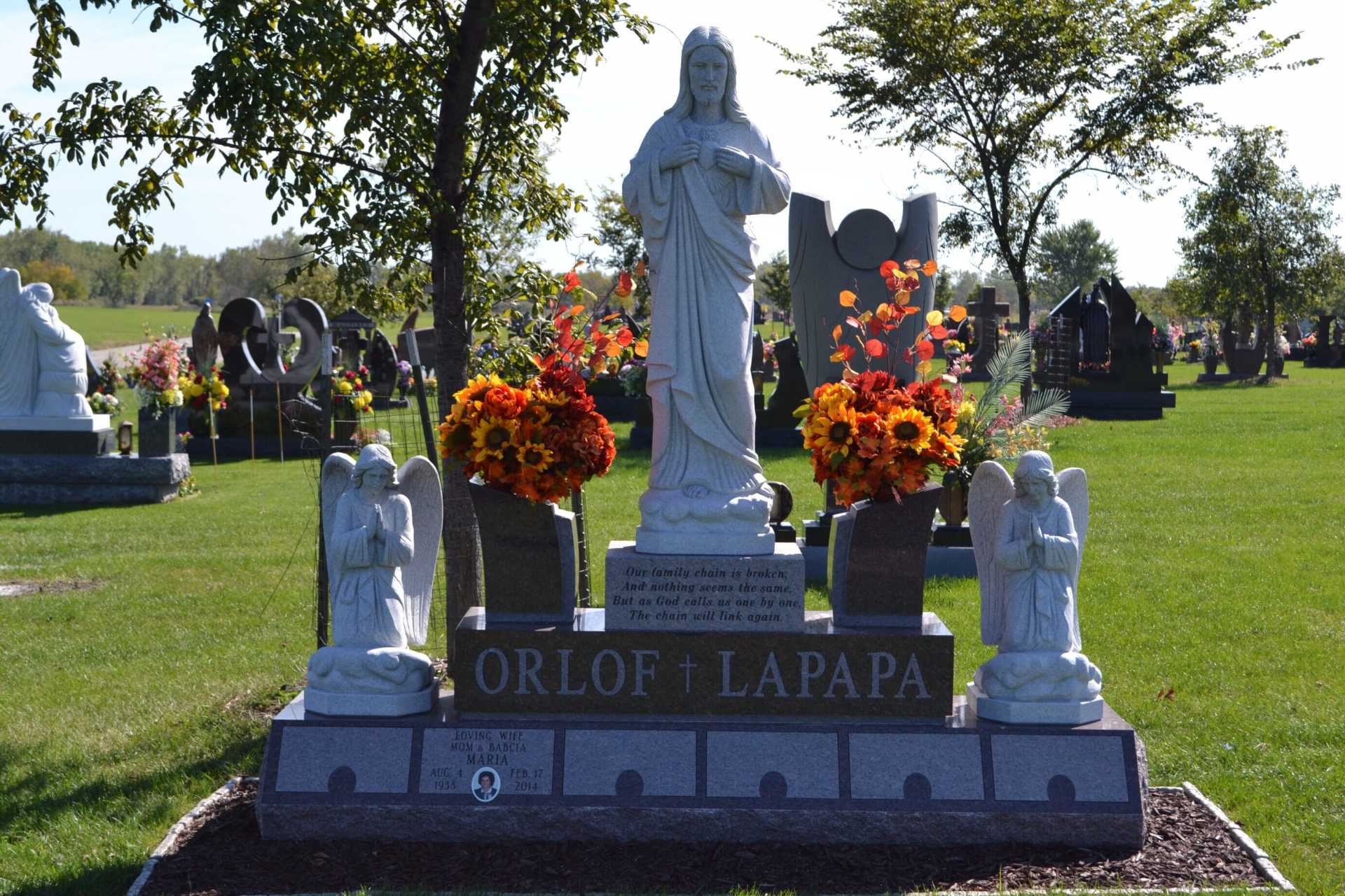 White statue of a woman in a cemetery, with colorful flowers, and the name 