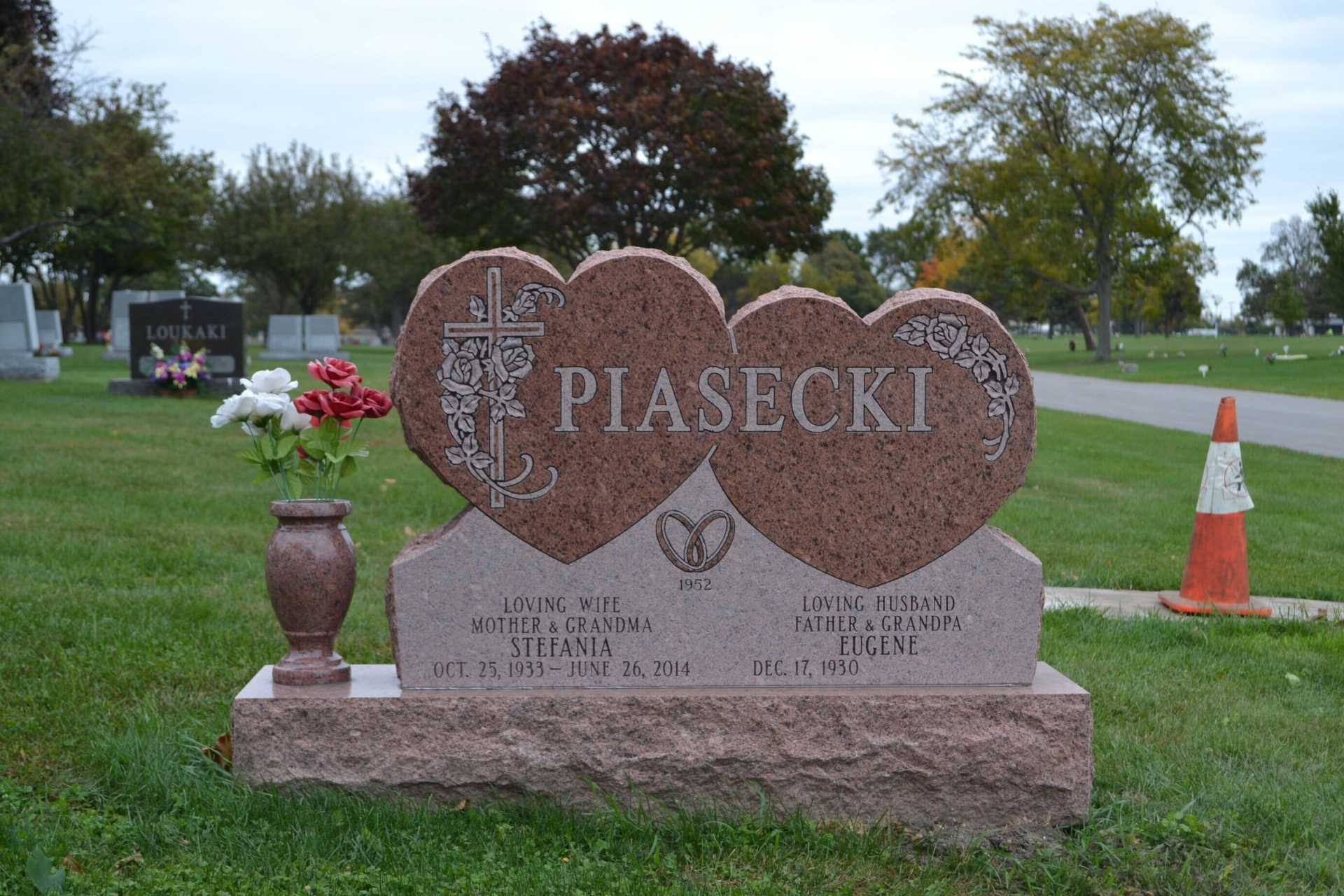 Grave marker with two heart-shaped sections for the Piasecki family, red granite, with a vase of flowers.