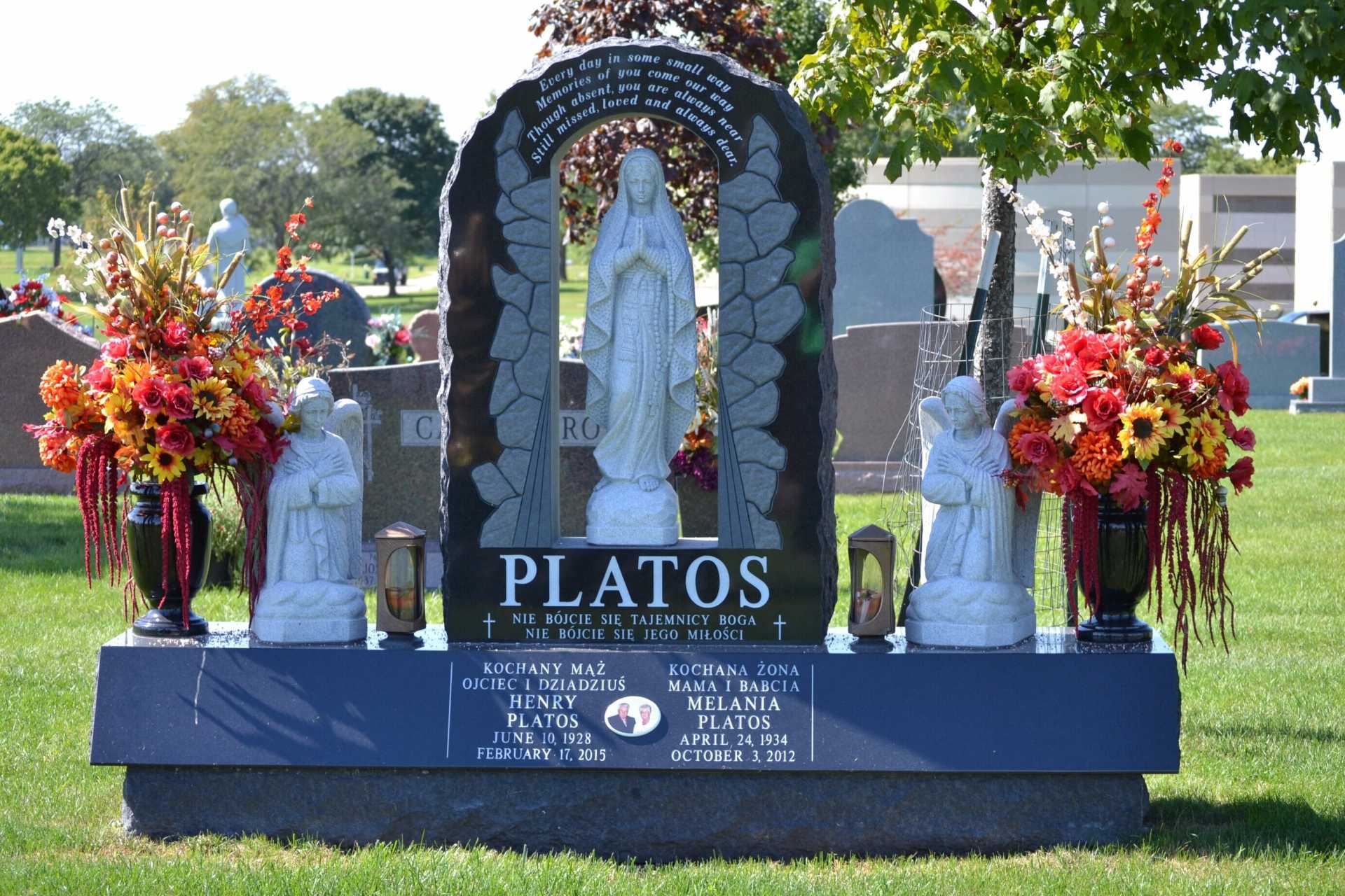 Monument in a cemetery, featuring a Virgin Mary statue flanked by floral arrangements and angel statues, green lawn.