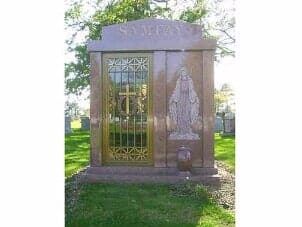 Granite mausoleum with ornate gold door, sculpted statue of Virgin Mary, and green grass.
