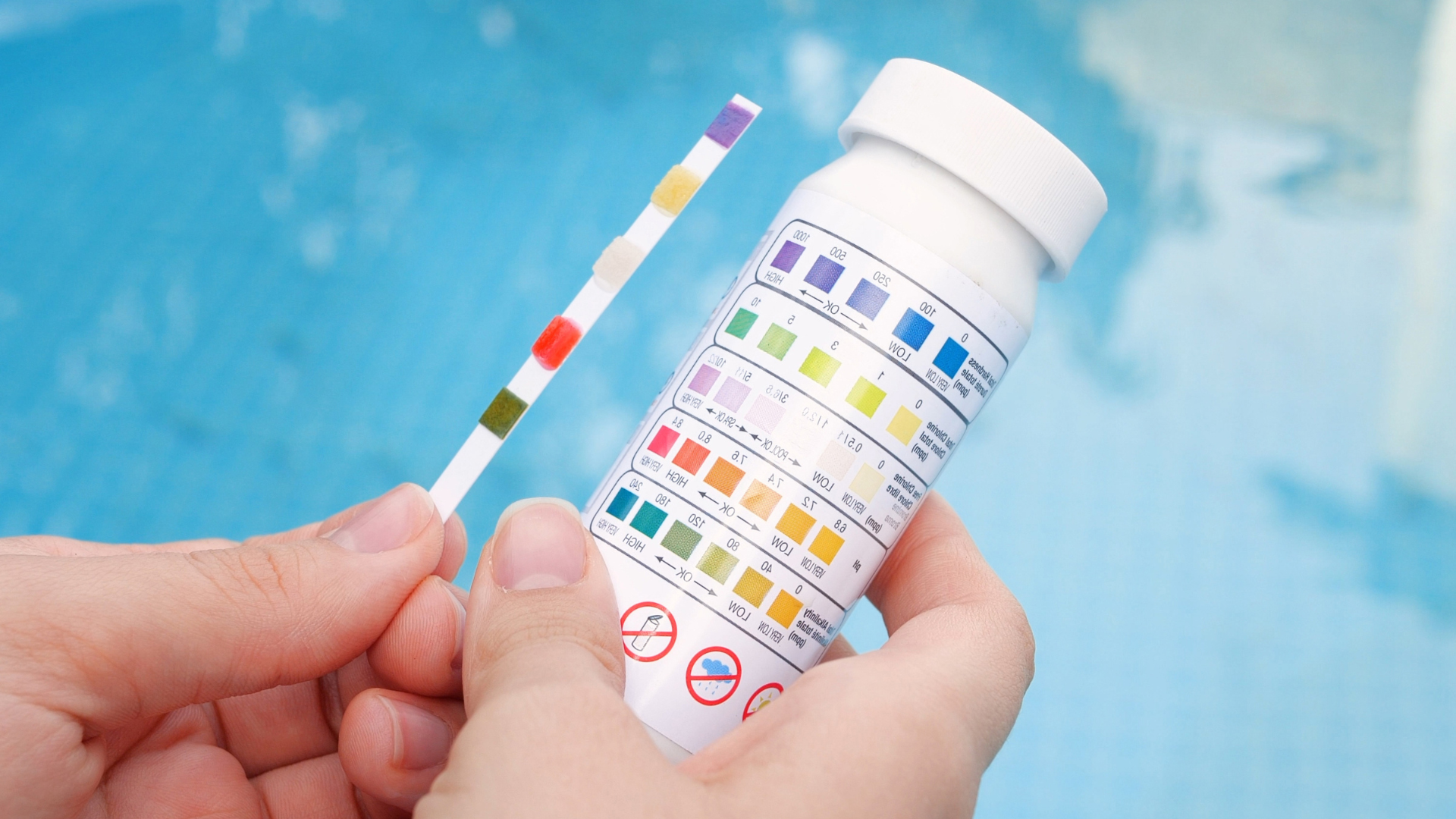 Hands holding a pool test strip next to a matching color chart bottle against a blurred blue water background.
