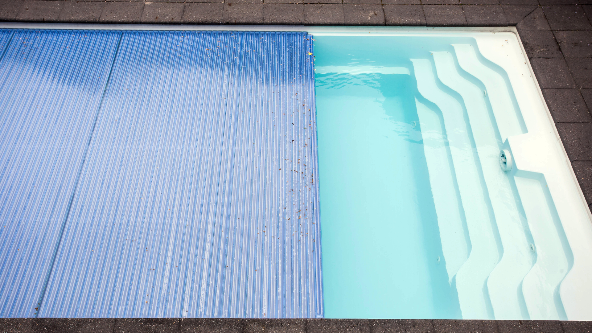A rectangular swimming pool partially covered with a blue bubble cover, revealing white stepped seating at one end.