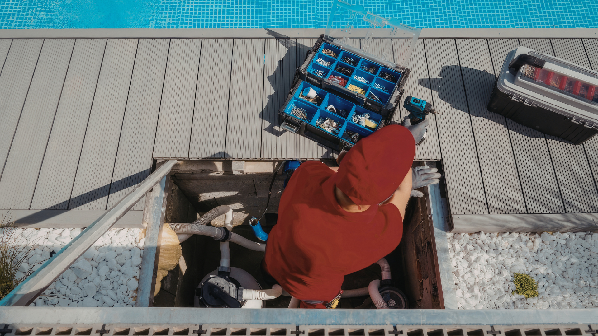 A technician in a red shirt repairs pool plumbing equipment inside an open utility pit on a pool deck.