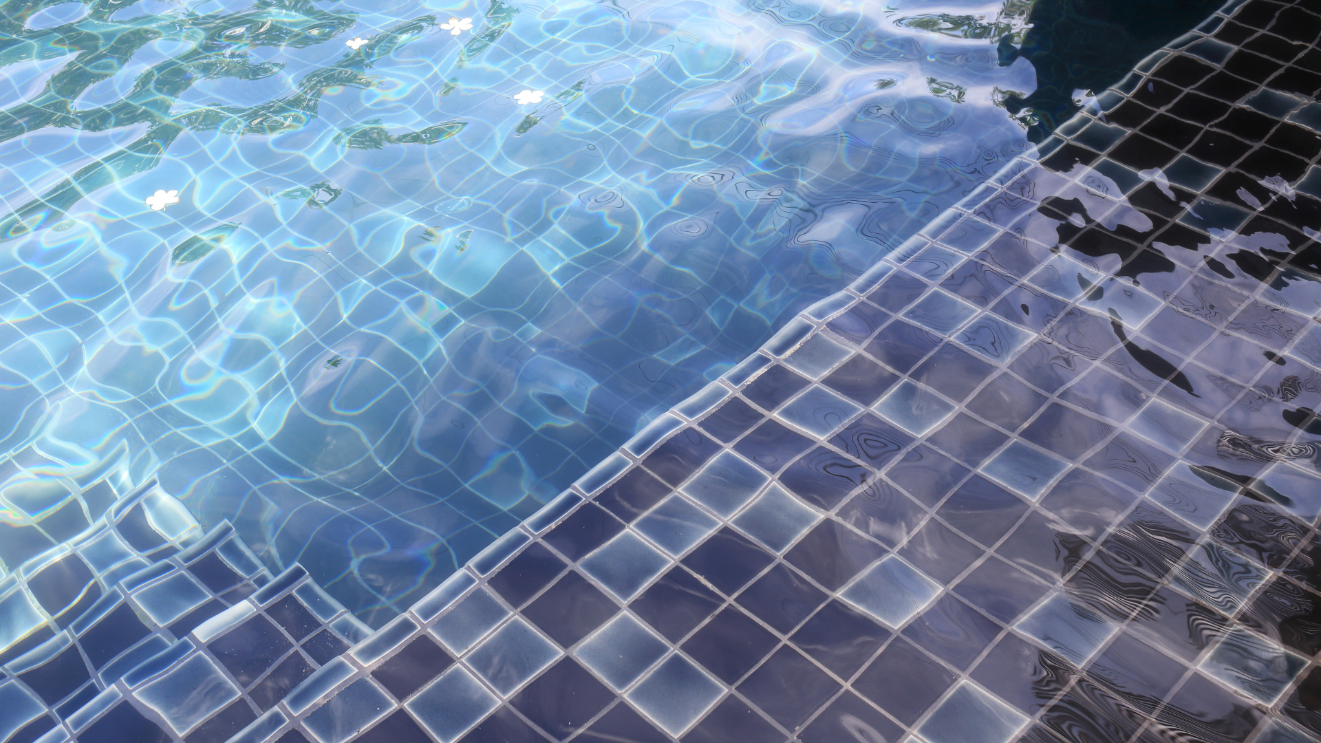 A close-up view of clear, rippling pool water over dark blue, square mosaic tiles on the steps and pool floor.