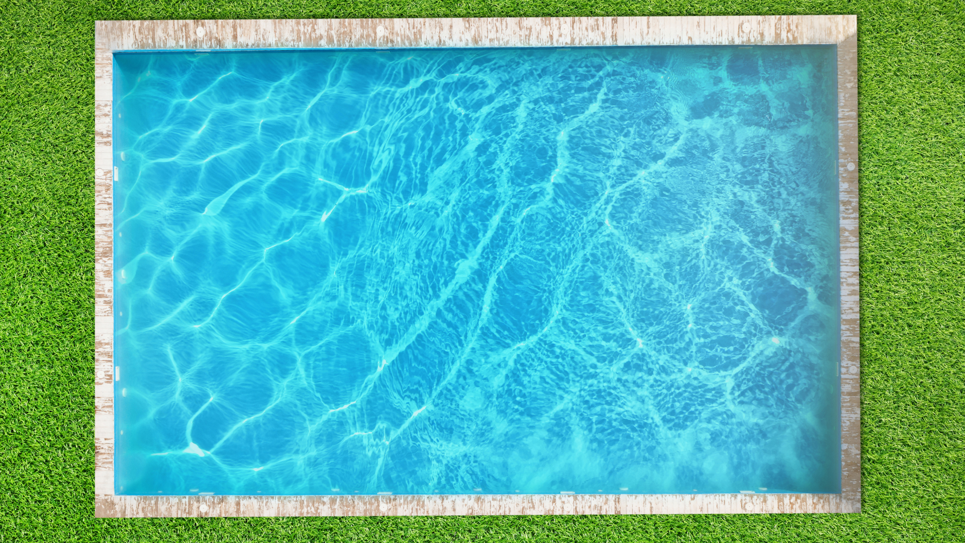 A rectangular swimming pool with blue, rippling water surrounded by a stone border and green grass, viewed from above.