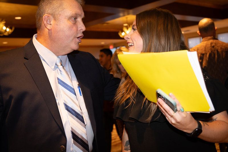 A man in a suit and tie is talking to a woman holding a yellow folder.