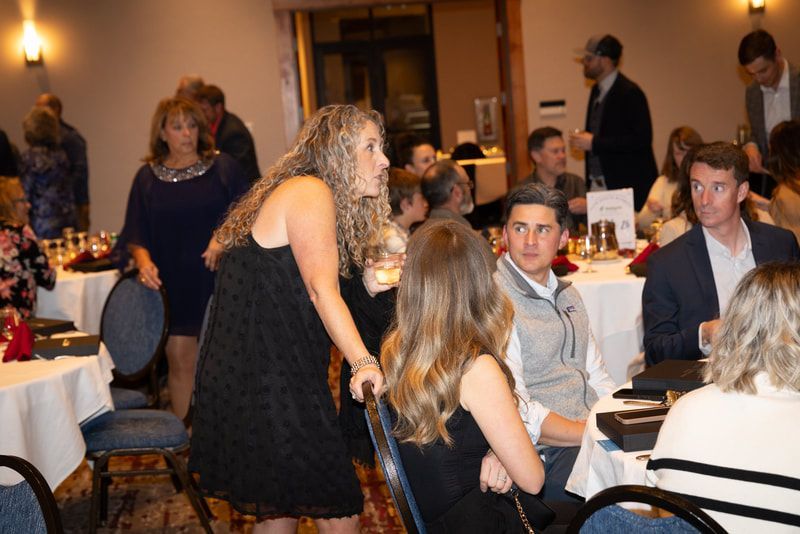 A woman in a black dress is standing in front of a group of people sitting at tables.