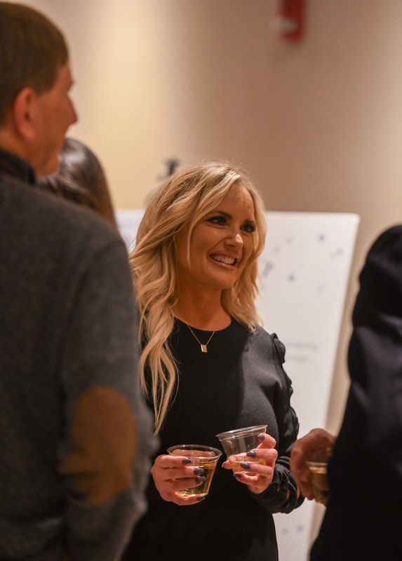 A woman in a black dress is holding two glasses of wine and smiling.