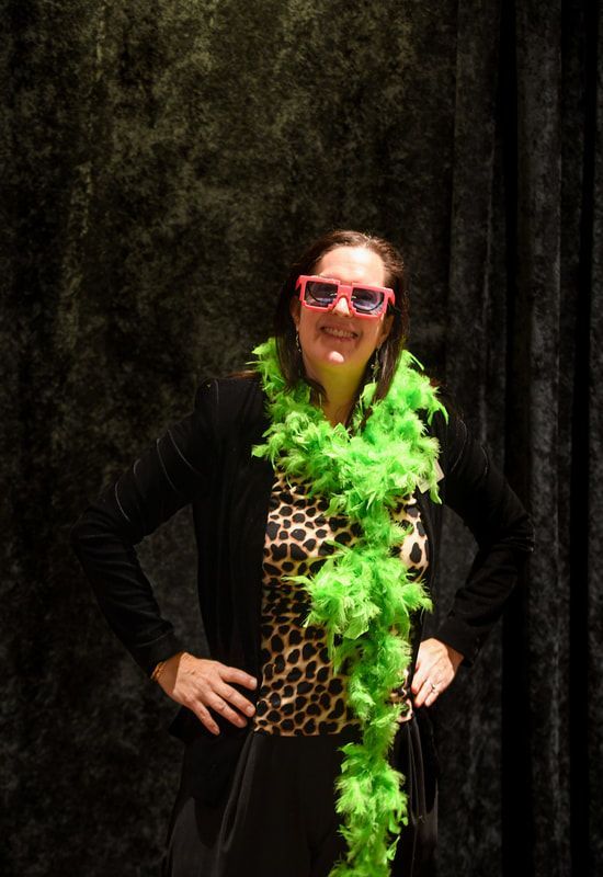 A woman wearing sunglasses and a green boa is standing in front of a black curtain.