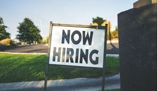 A now hiring sign is sitting on the side of the road.
