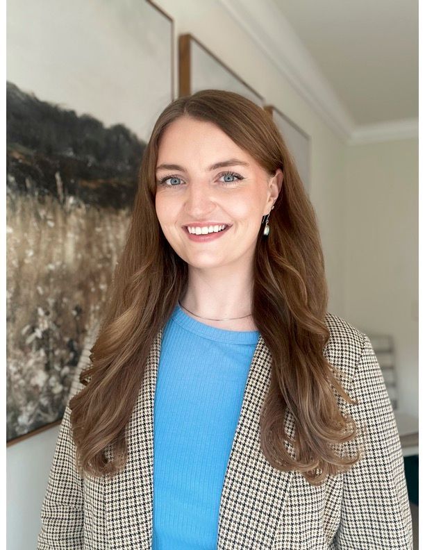A woman in a blue shirt and a plaid jacket is smiling in front of a painting.