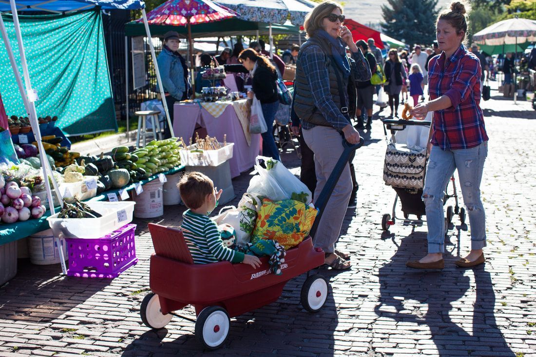A little girl is sitting in a red wagon at a farmers market.