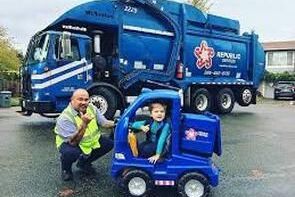 A man is kneeling next to a garbage truck and a child is sitting in a toy truck.