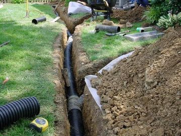 Trenched yard with corrugated drainage pipe being installed beside a grassy lawn.