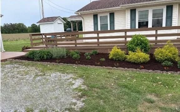 Front yard with a wooden ramp and railings leading to a house, bordered by shrubs and mulch.