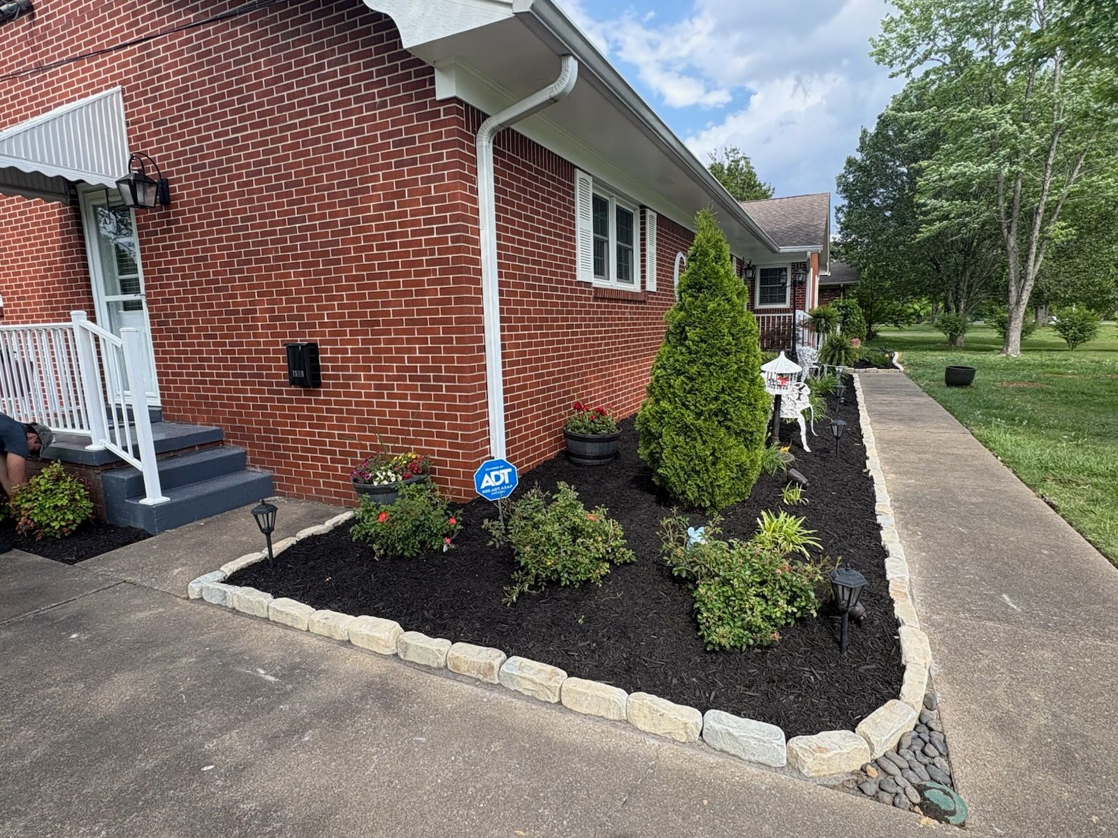 Brick house exterior with mulched flower bed, small shrubs, and a concrete walkway along the side