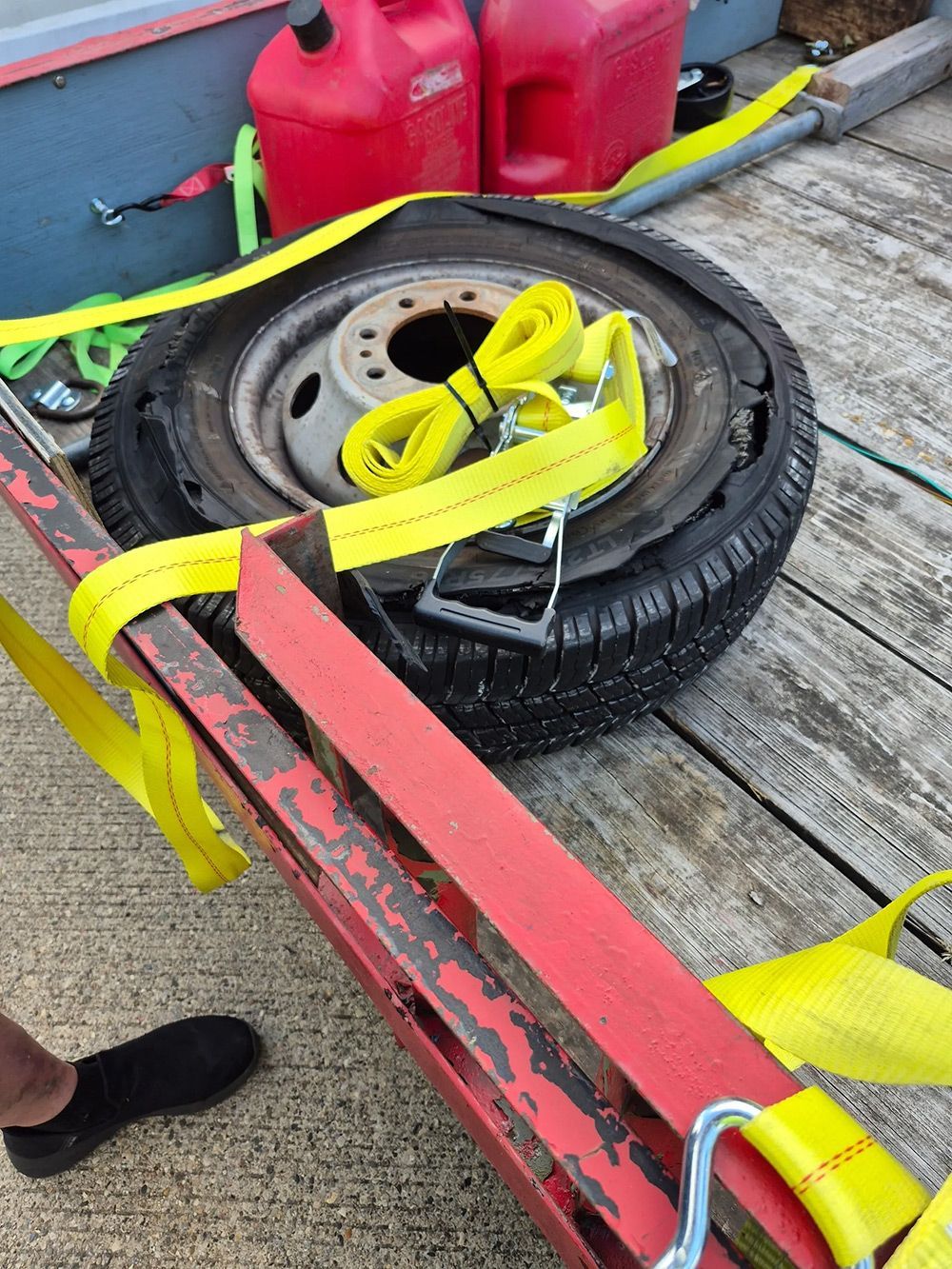 Flat tire secured to a red trailer with yellow straps, setting outdoors near gas cans.