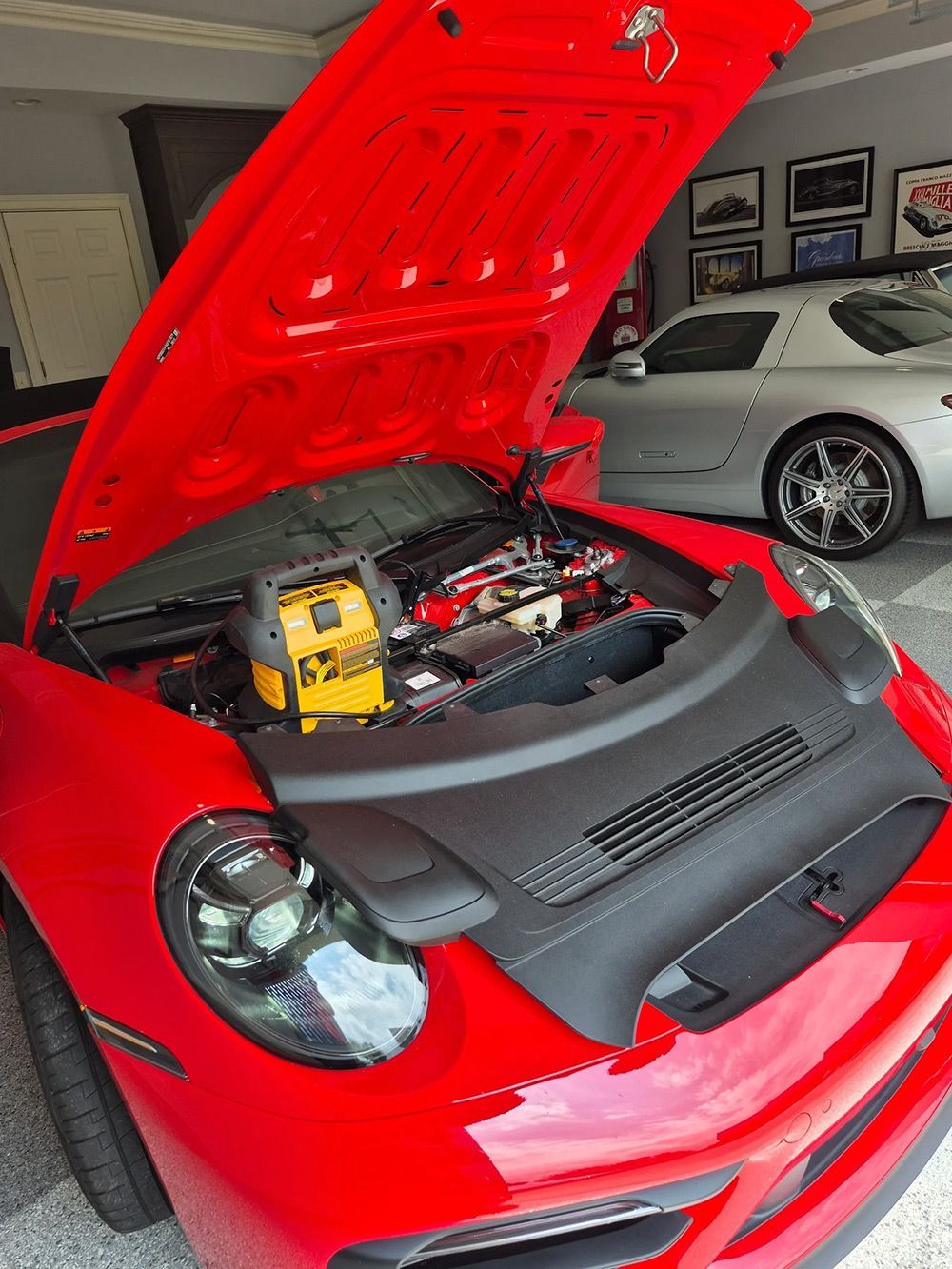 Red Porsche with open hood, jump starter attached, garage setting, and silver car in background.