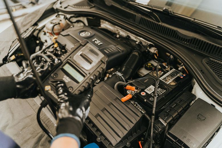 Mechanic with gloves working on a car engine, using a diagnostic tool. Focus on black battery and TSi engine components.