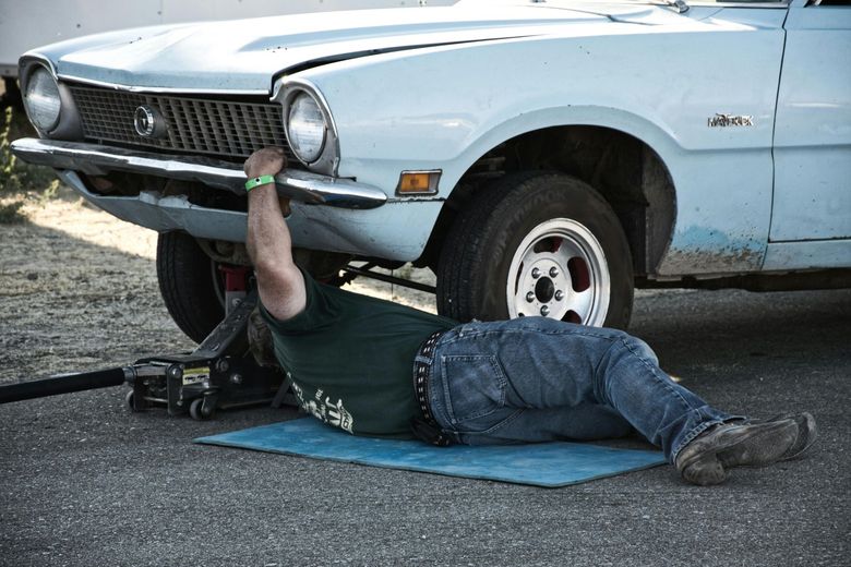 Man working on a light blue classic car, lying on a mat, jacked up on a paved surface.