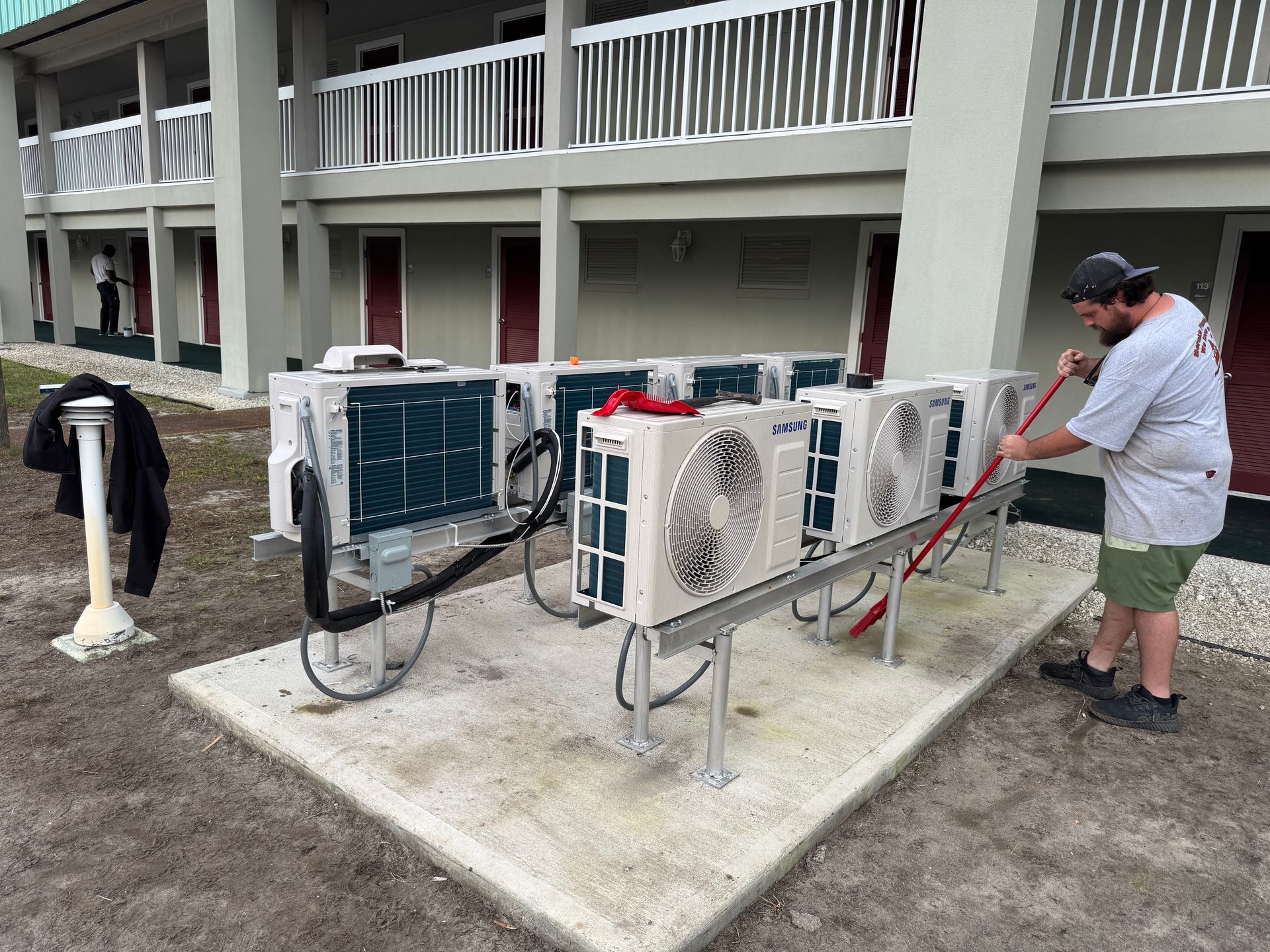 Man cleaning air conditioning units outside a light-colored building.