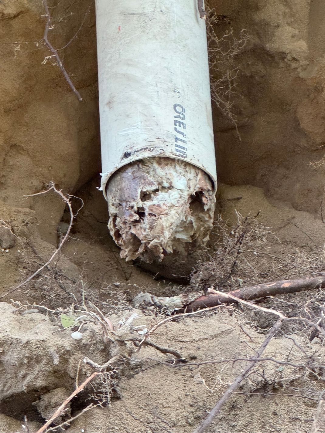 White PVC pipe damaged and exposed in a dirt trench, showing deterioration at the lower end.