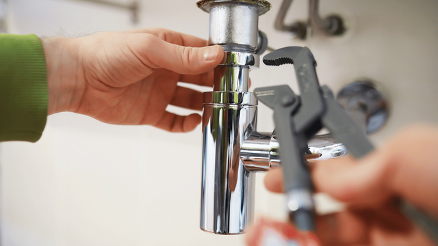 Hands of a person using a wrench to adjust a chrome pipe under a sink.