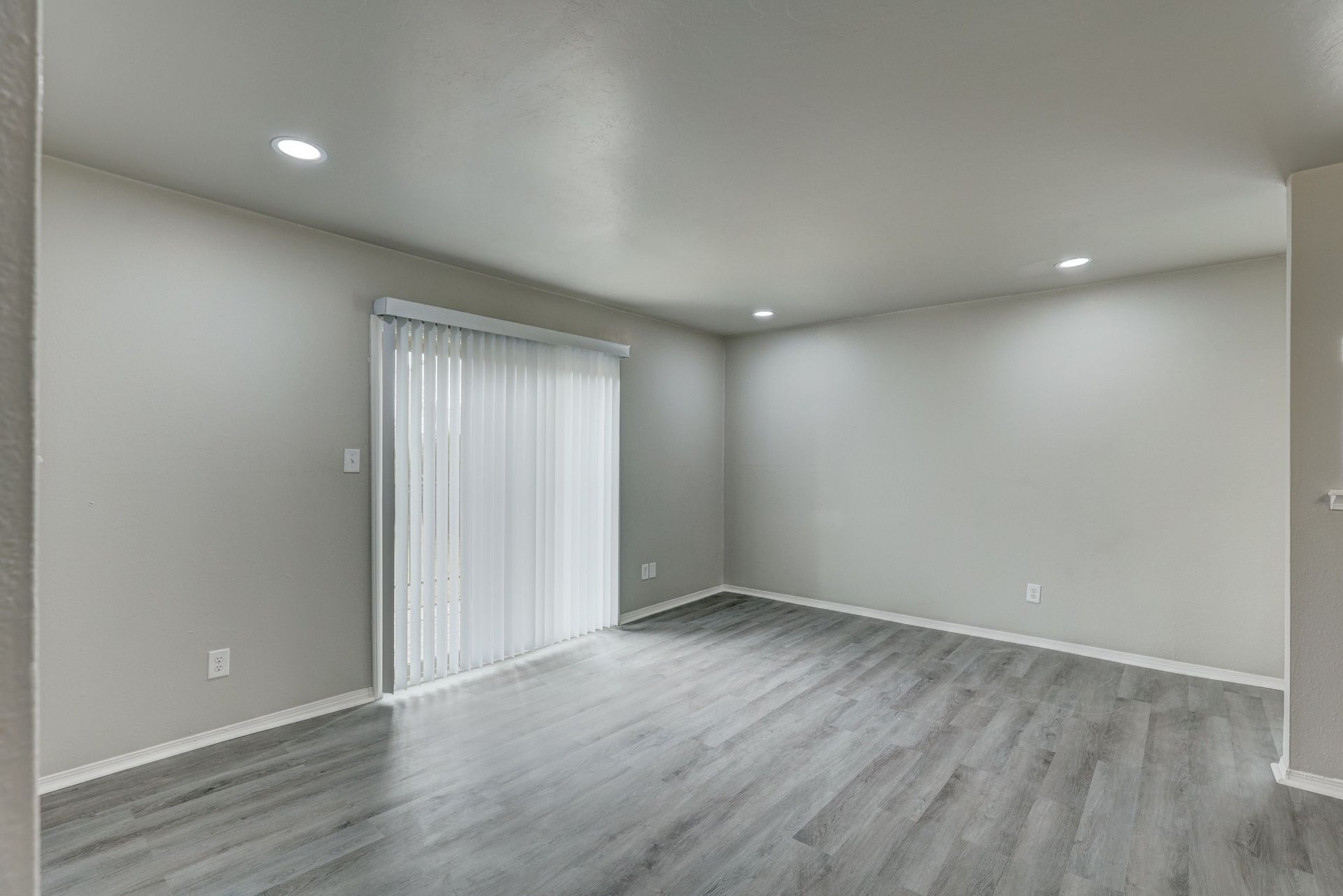 Empty living room with gray walls and flooring, sliding glass door, and recessed lighting.