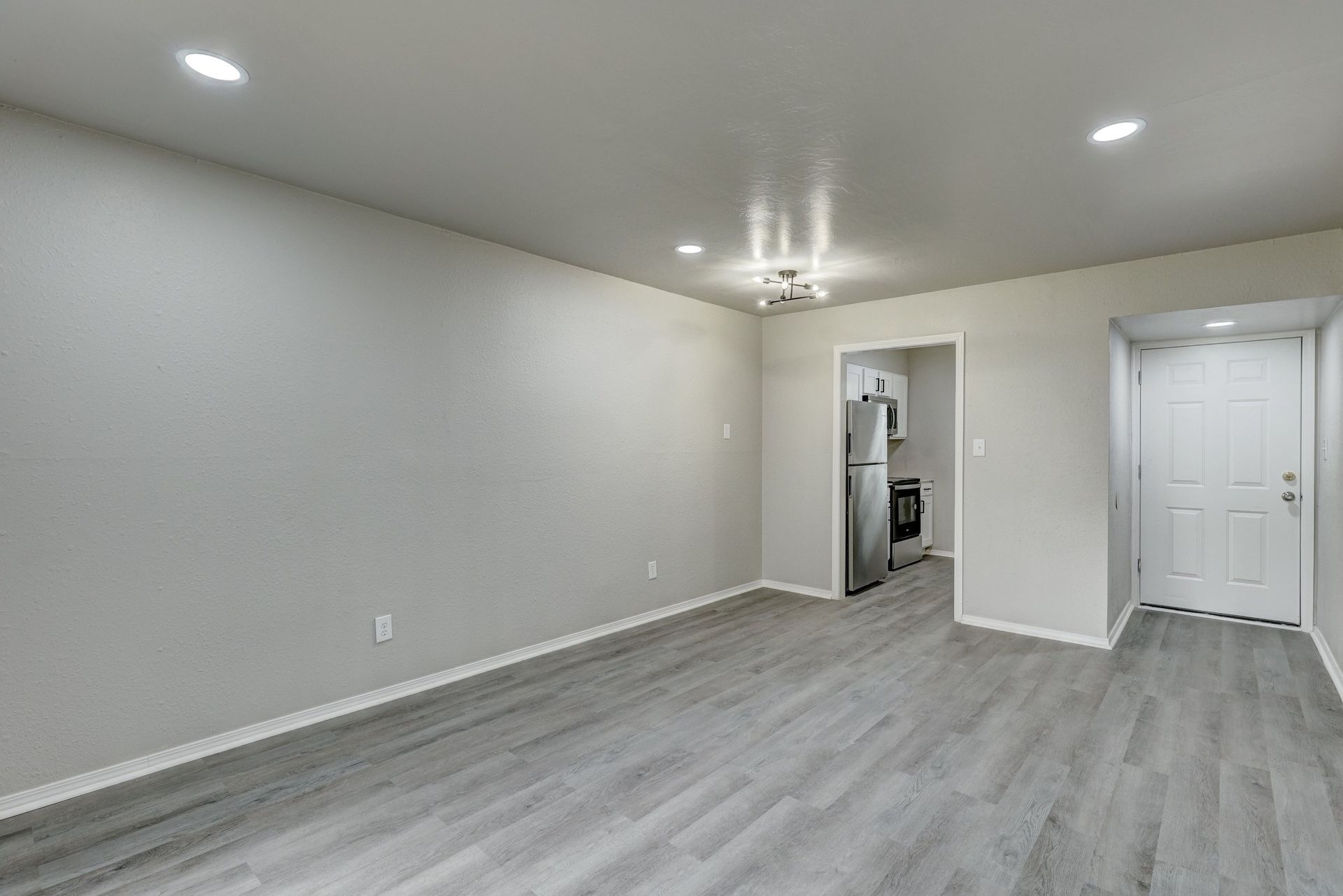 Empty living area with gray walls and flooring, leading to a kitchen with appliances.