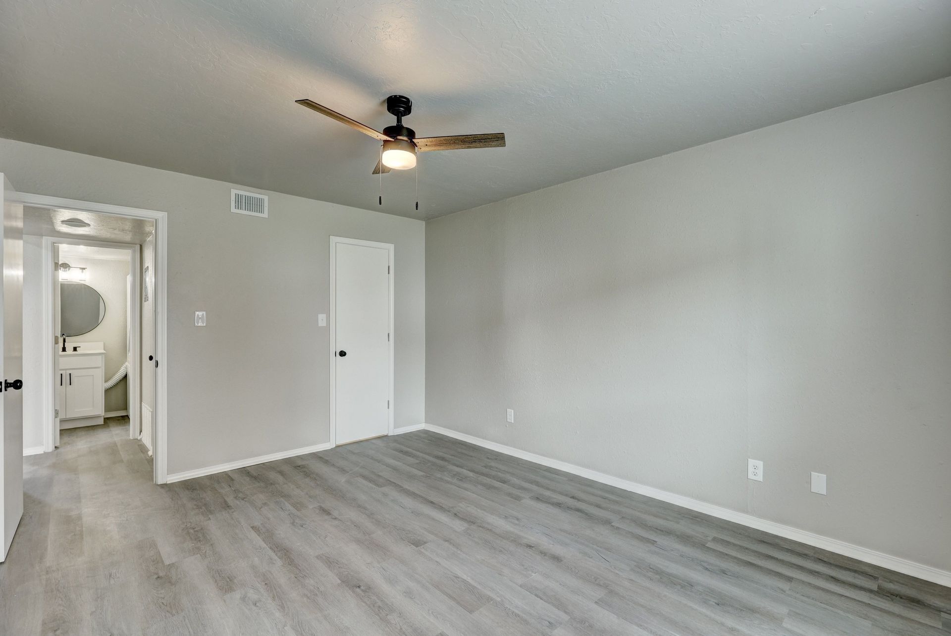 Bedroom with gray walls, wood-look floors, a ceiling fan, and an open doorway to a bathroom.