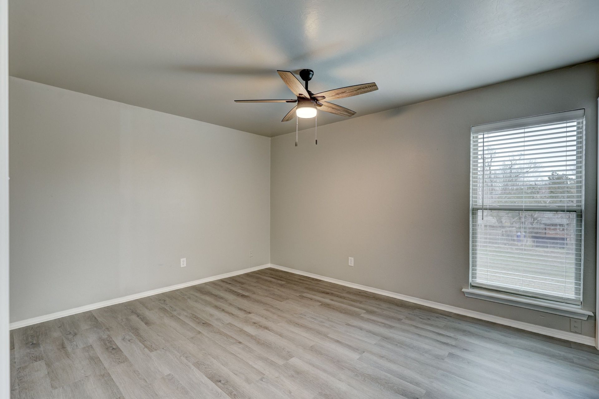Empty room with gray walls, wood-look floors, ceiling fan, and window with blinds.
