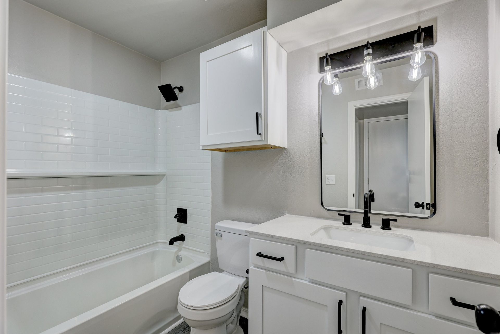 White bathroom with tub, toilet, vanity, and overhead cabinet; black fixtures.