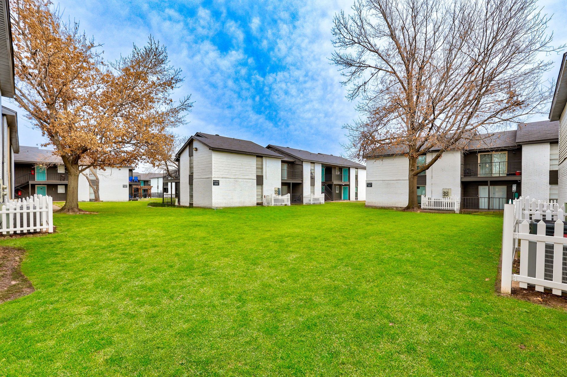 Apartment complex with white buildings, green grass, and bare trees under a blue sky.