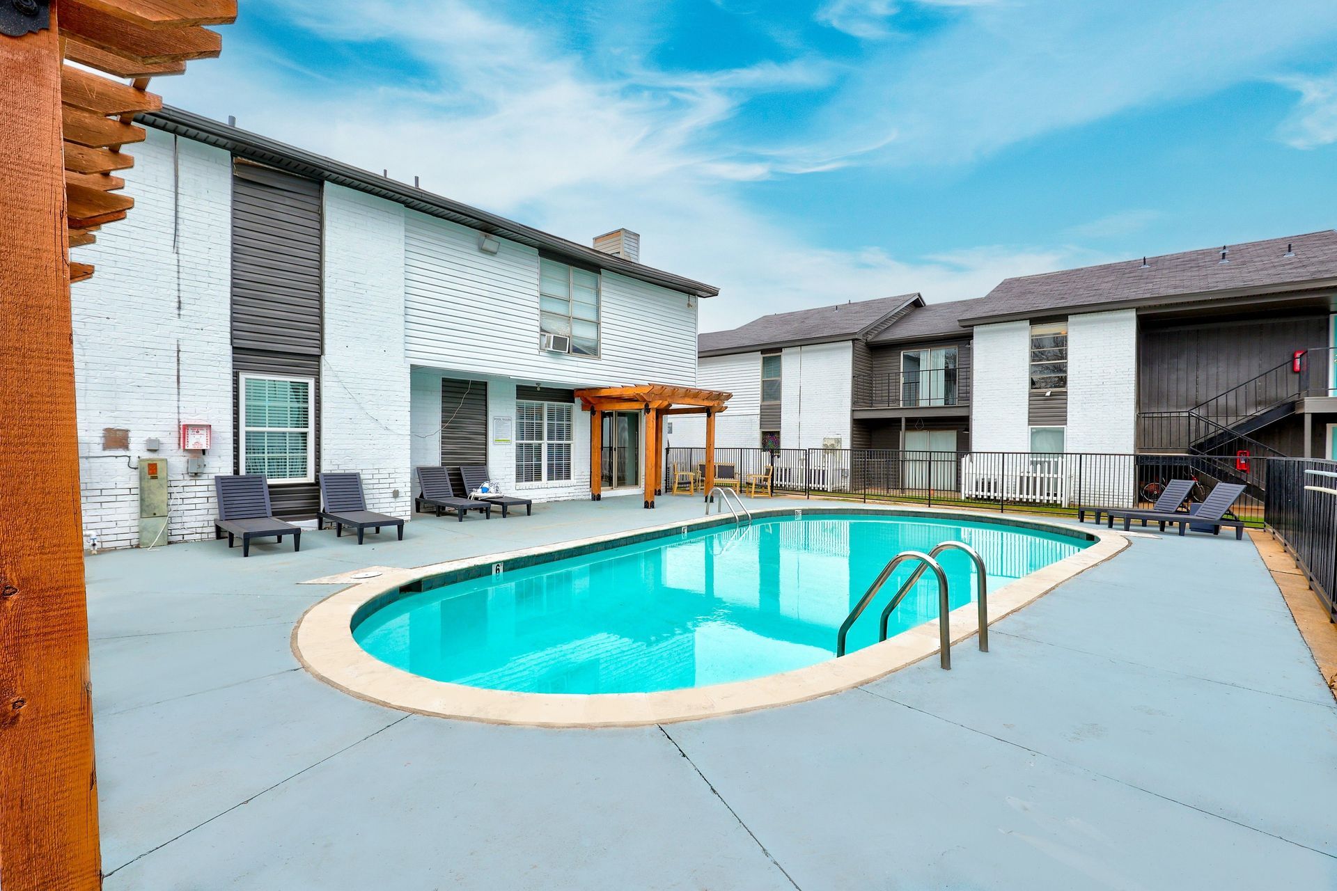 Apartment complex with a pool. Blue water, grey patio, and lounge chairs. Cloudy sky.