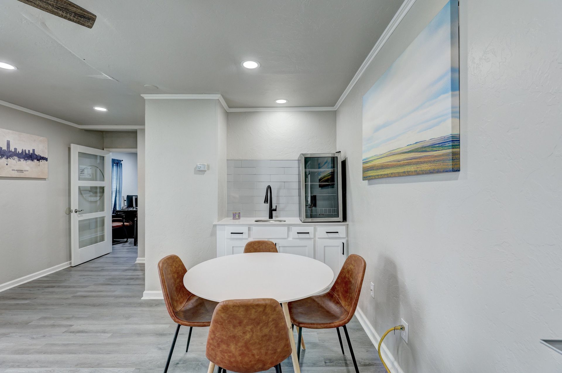 Dining area: Round table with four brown chairs. Mini-fridge and artwork on the wall.