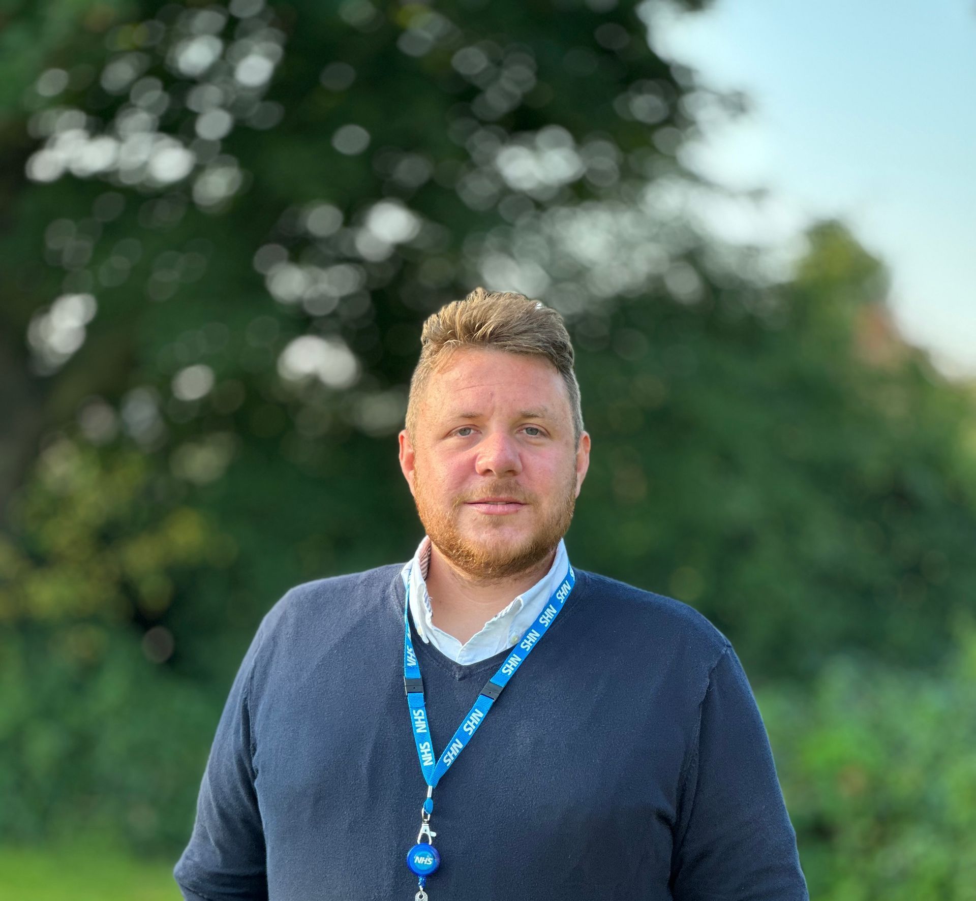 A man with brown hair and beard smiling with a blurred background. He is wearing a blue jumper over a white shirt and has an NHS lanyard around his neck. 