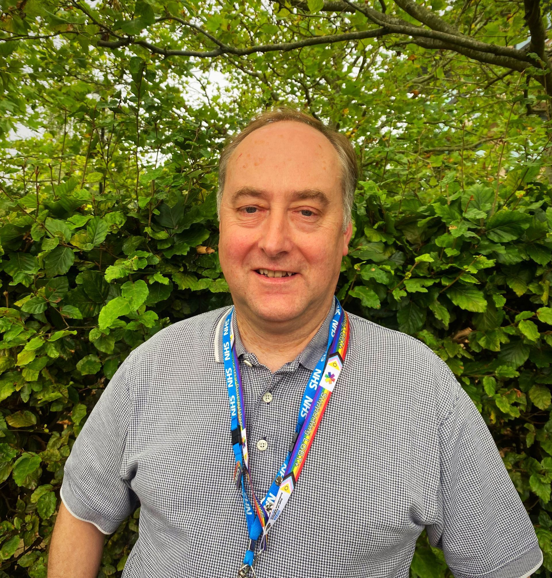 Man in blue shirt with a patterned tie. He is smiling and has a blue NHS lanyard on. 