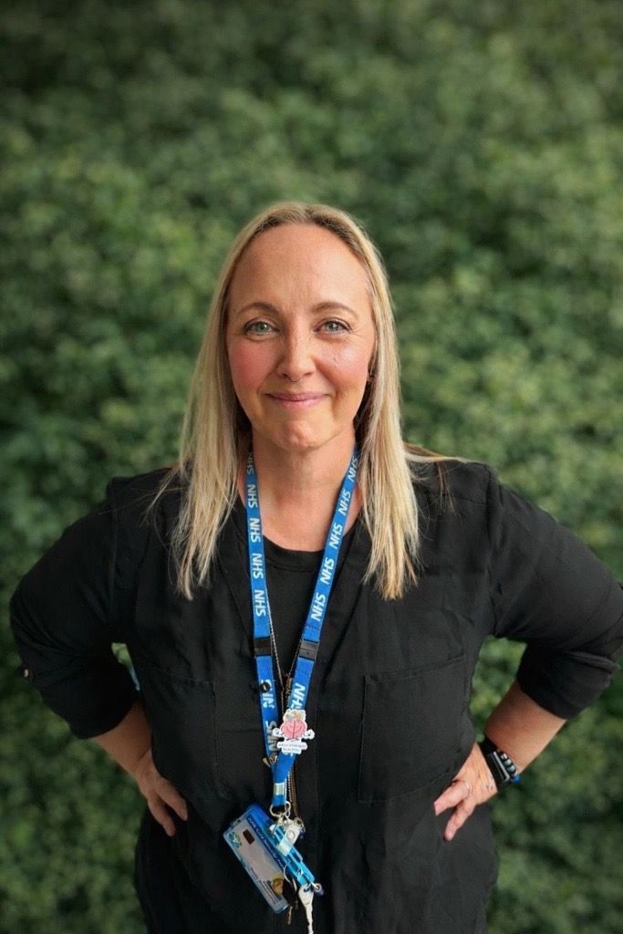 A lady with blonde hair smiling with a blurred background. She is wearing a white top, long necklace and black blazer.