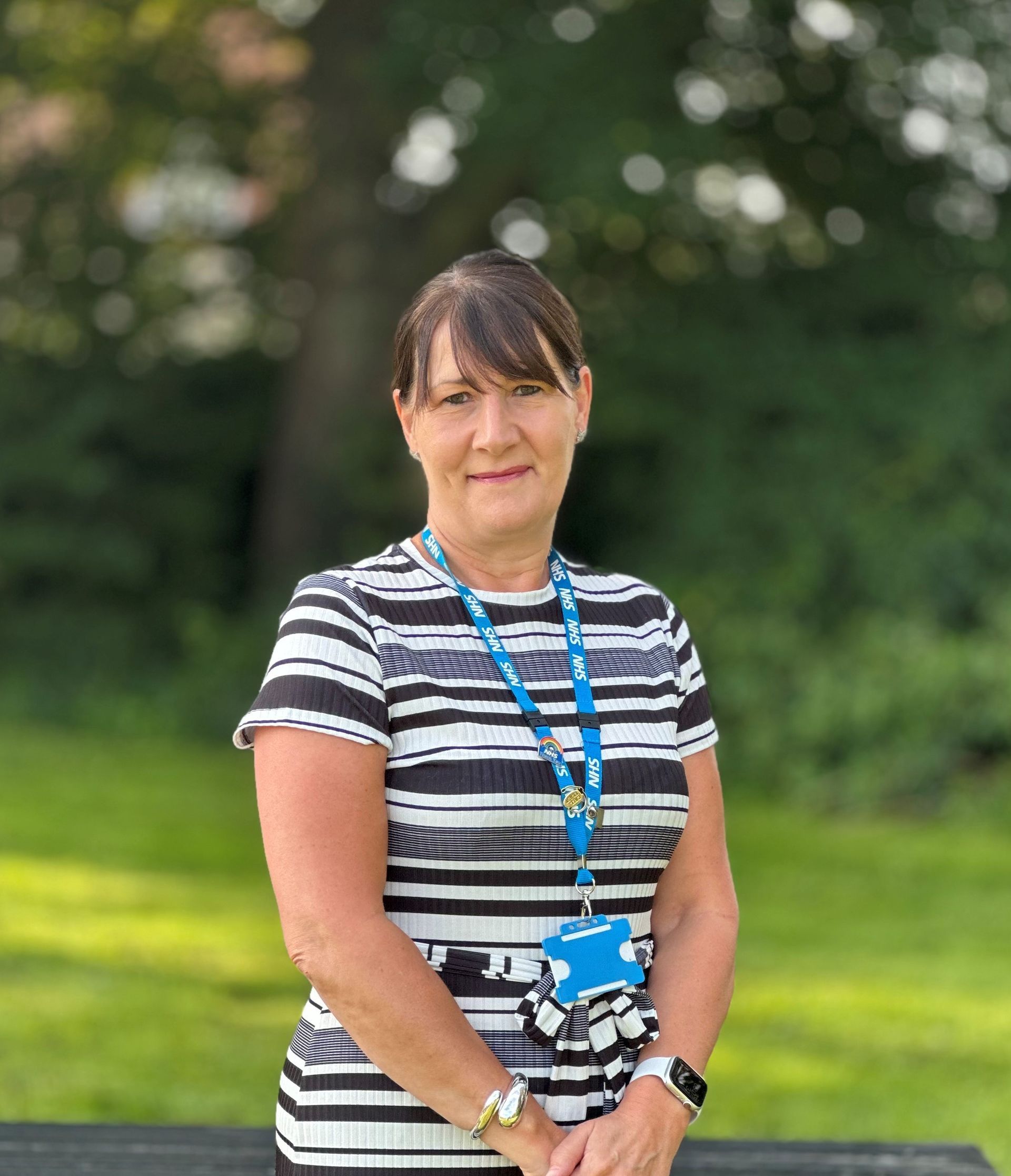 A lady with brown hair and a fringe smiling with a blurred background. She is wearing a black and white striped dress, her NHS lanyard and has her hands by her side. 