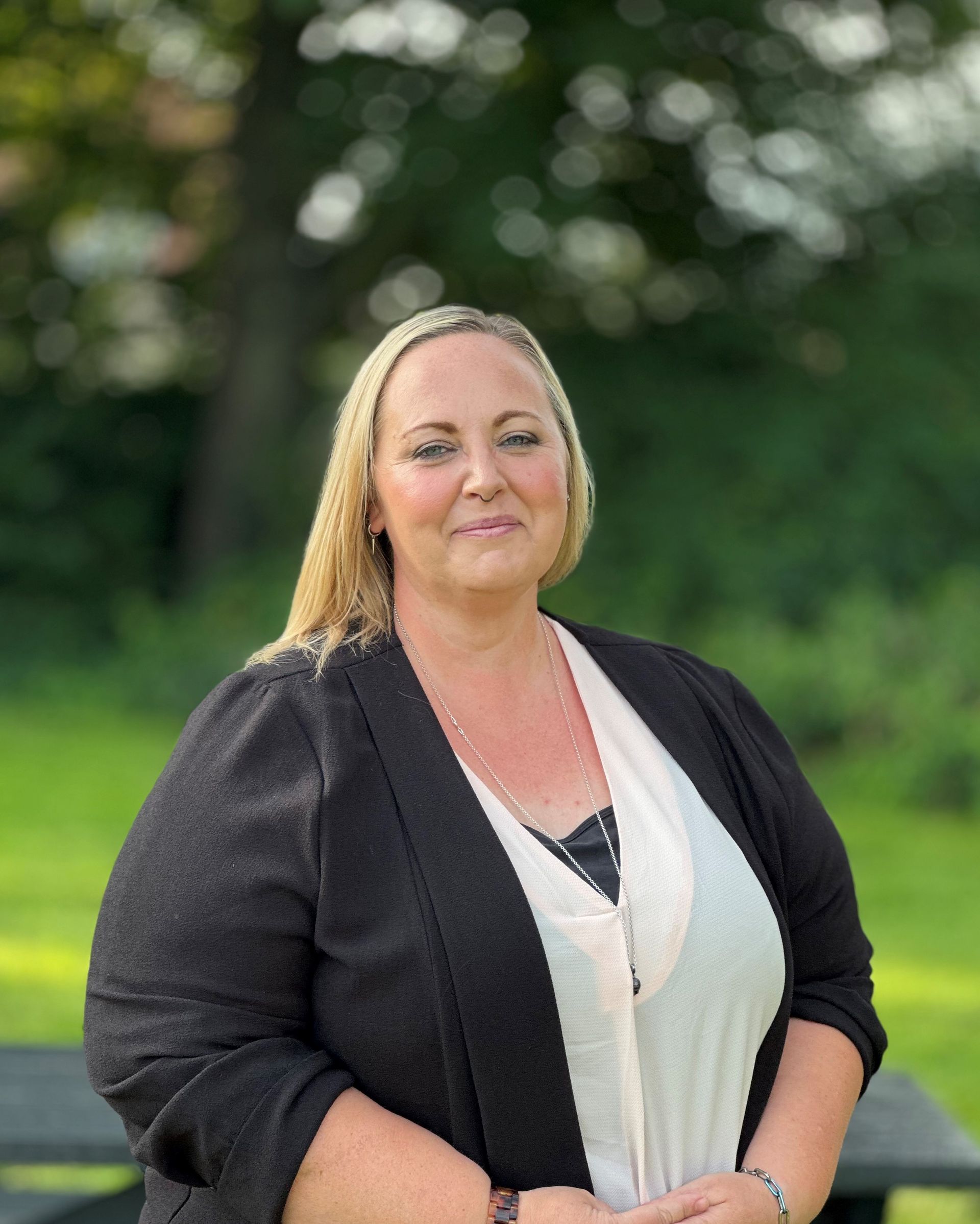 A lady with blonde hair smiling with a blurred background. She is wearing a white top, long necklace and black blazer.