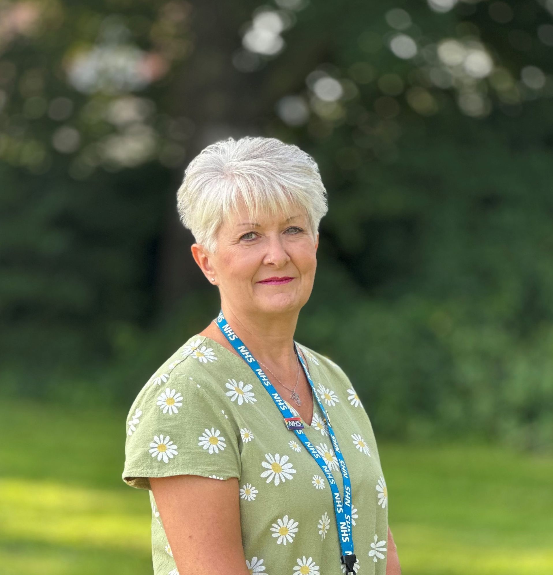 Sally, a member of CallEEAST is wearing a light green dress with white and yellow daisies on. She has short white hair and is wearing pink lipstick. She has her hands by her sides, is wearing an NHS lanyard and a silver necklace and is smiling. The background of trees is blurred.
