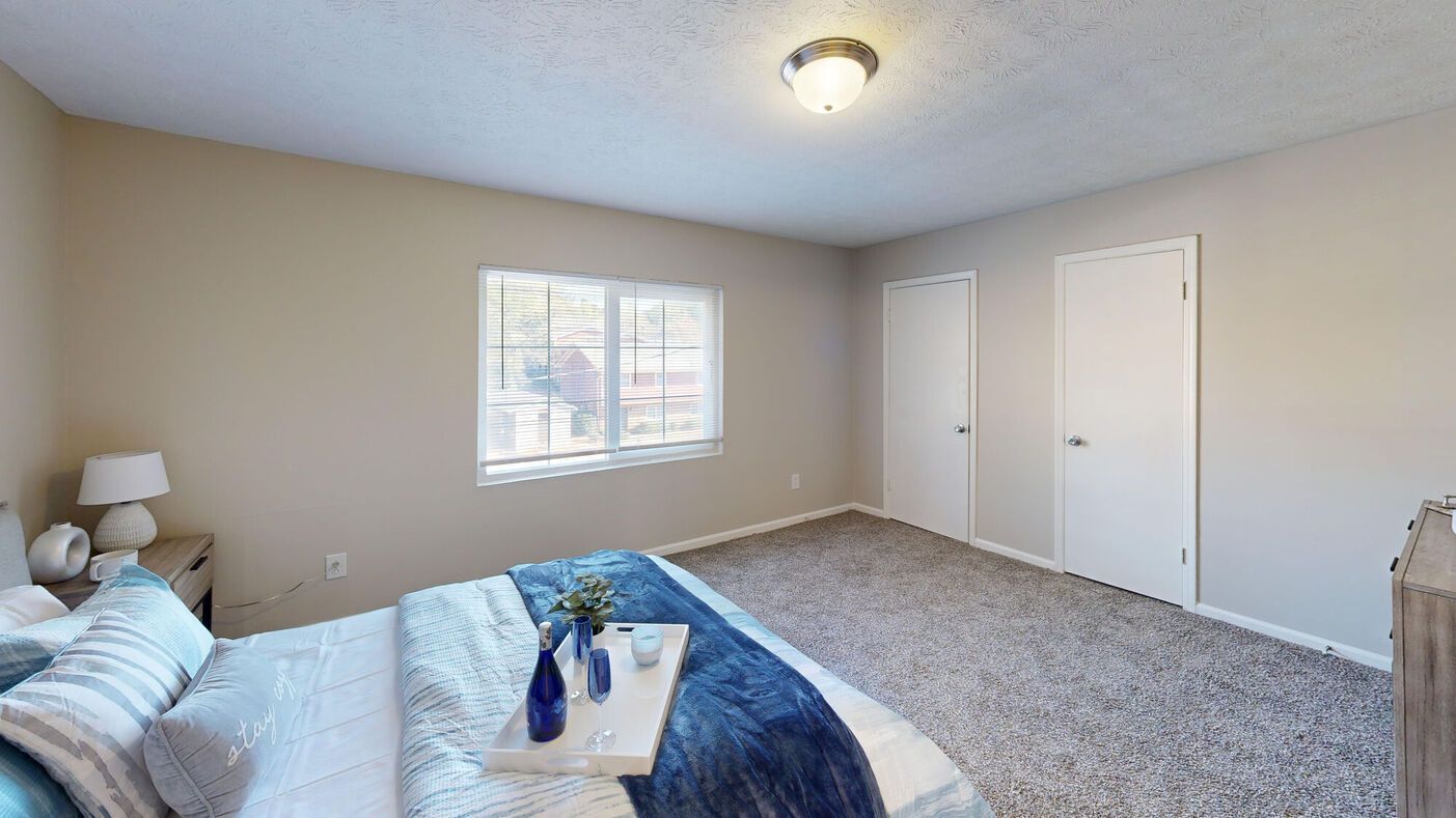 Bedroom with bed, window, two doors, beige walls, and gray carpet.