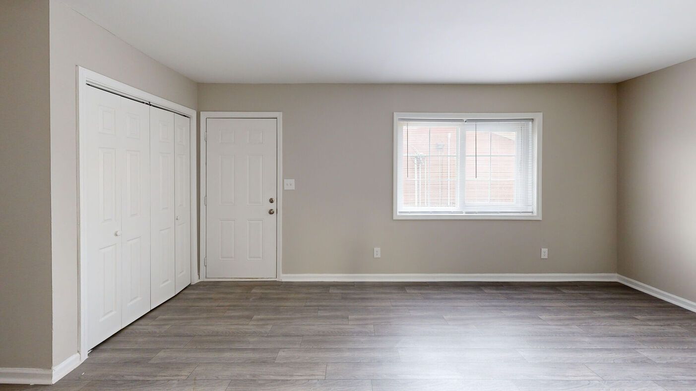 Empty room with white closet doors, a door, and a window with blinds; gray walls and wooden floor.
