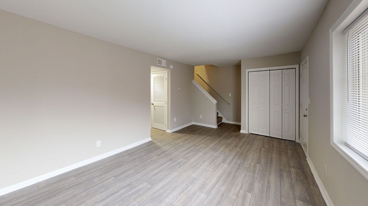 Interior view of a living space with light wood flooring, stairs, and a closet. Neutral walls and bright light.