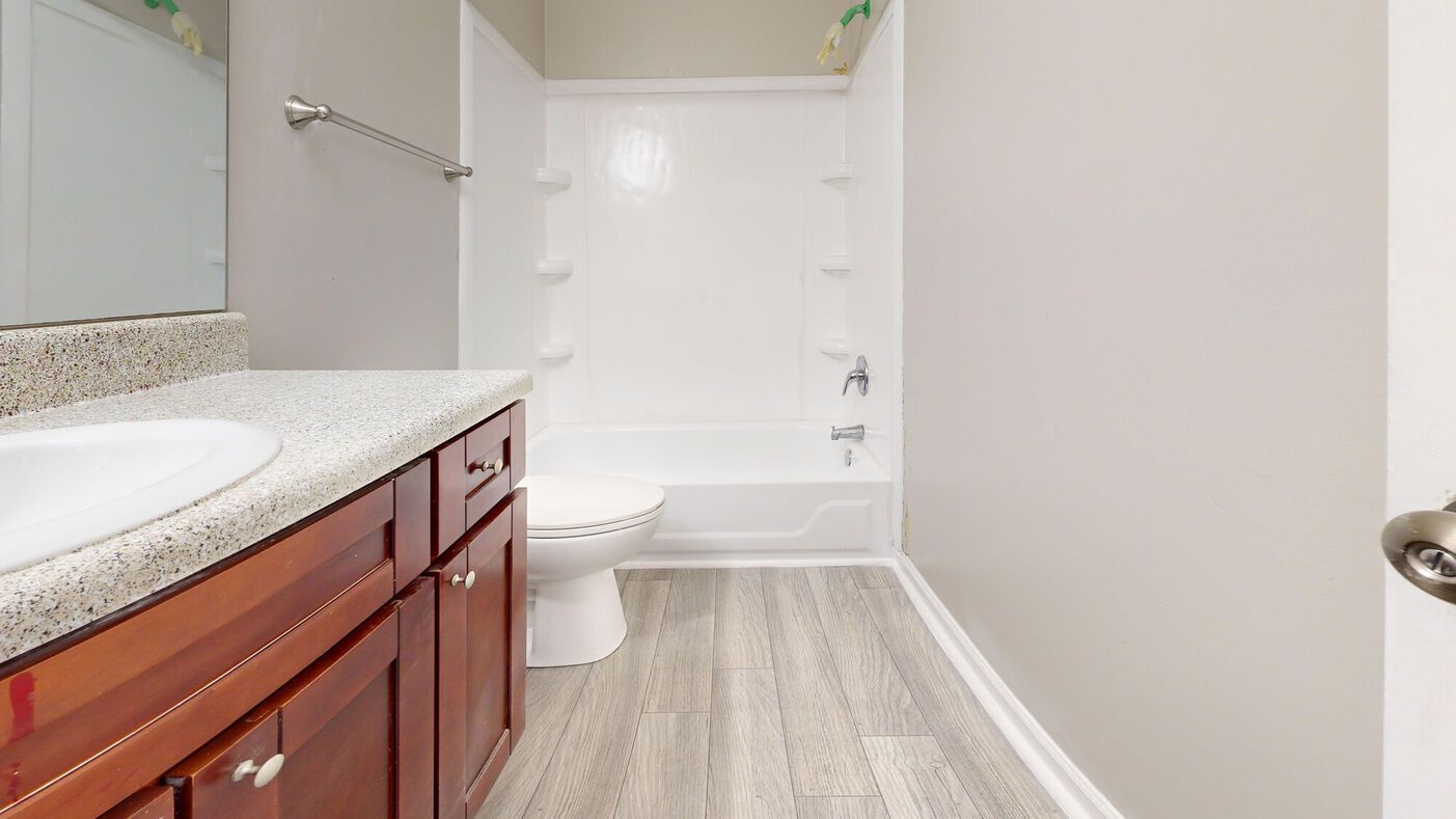 Bathroom with wood-toned vanity, white toilet and tub, and light gray walls and flooring.