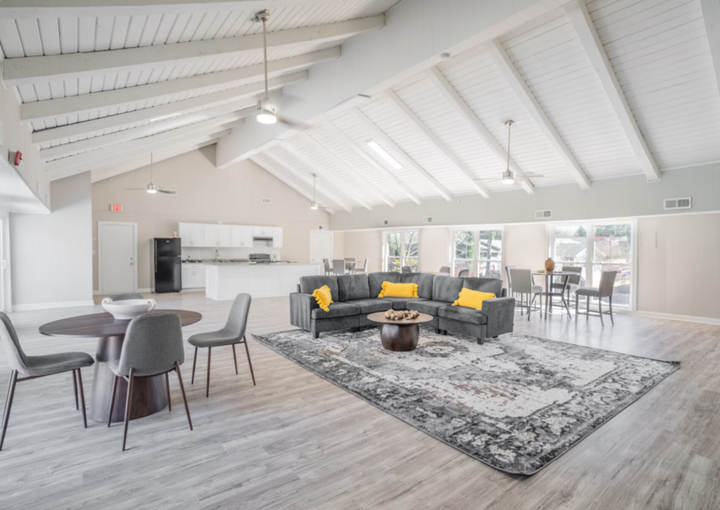 Spacious community room with gray sectional sofa, round tables, and white beamed ceiling.