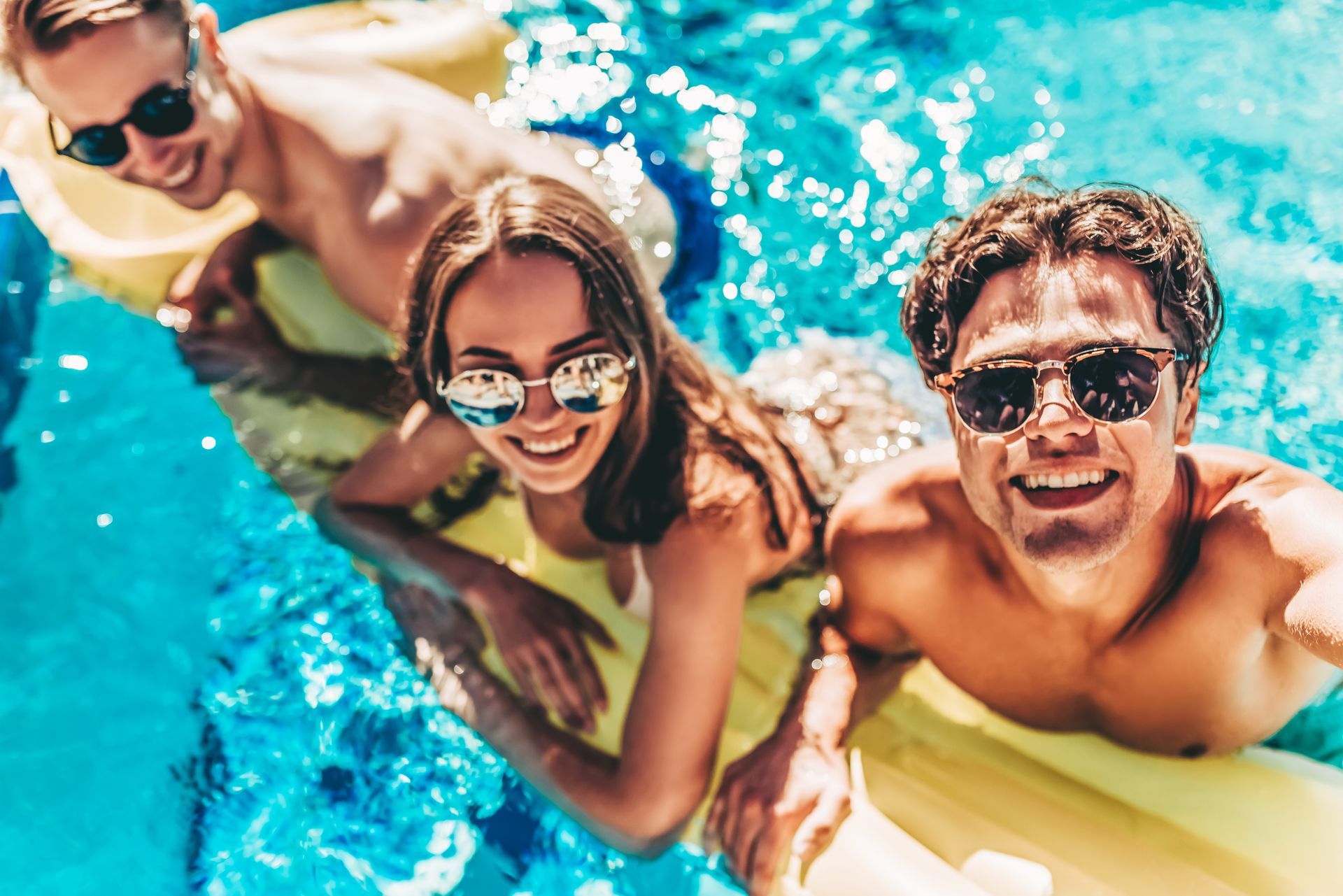 Three people in sunglasses smiling in a swimming pool on a yellow float.